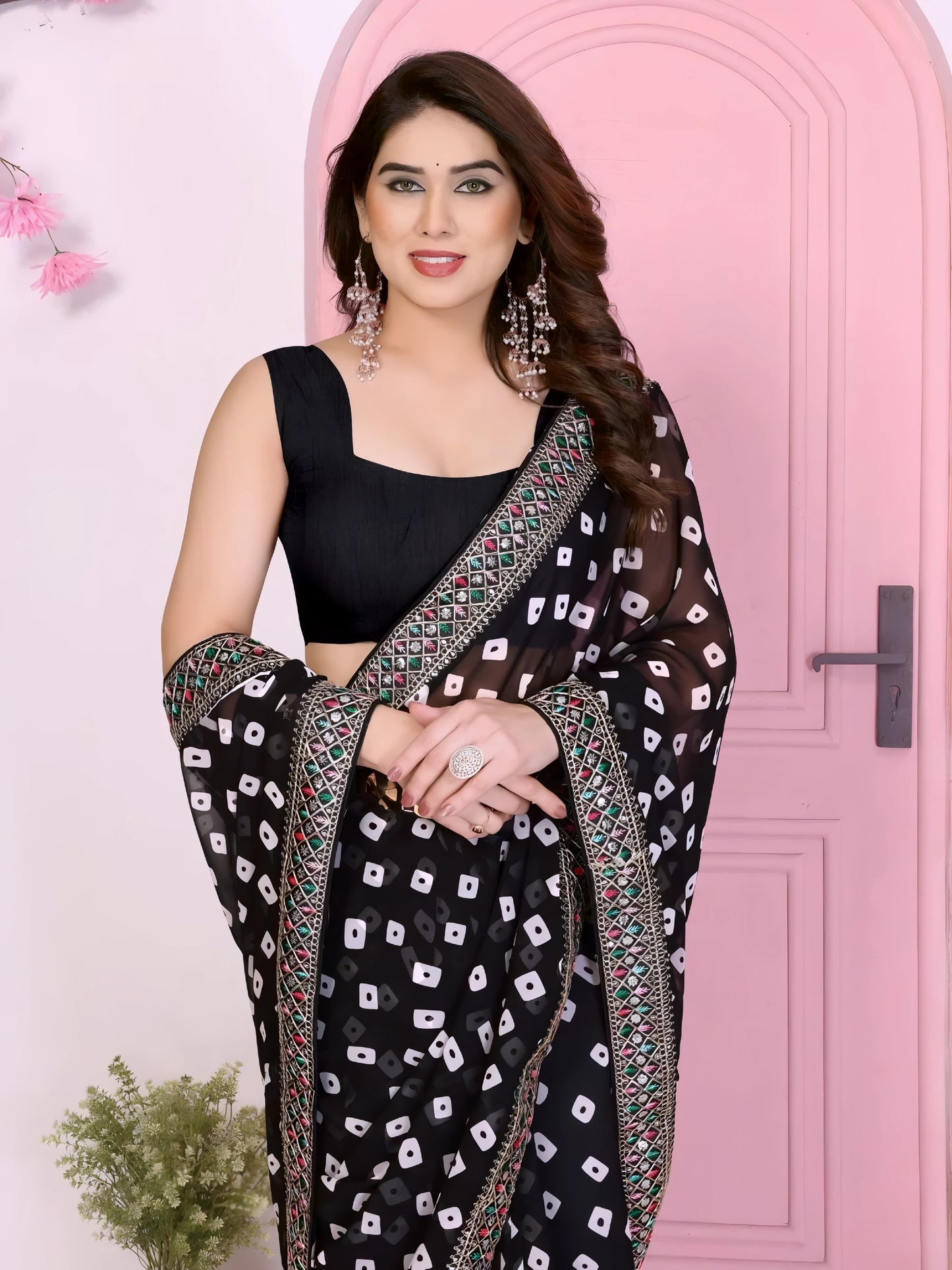 Woman wearing a vibrant pink bandhani saree with traditional white tie-dye patterns and a decorative embroidered border, paired with a sleeveless matching blouse. She stands gracefully in front of a pastel pink arched doorway, accessorized with statement earrings and bangles, surrounded by earthy clay pots, a small green foliage vase, and pink decorative branches in a contemporary minimalist studio setting.