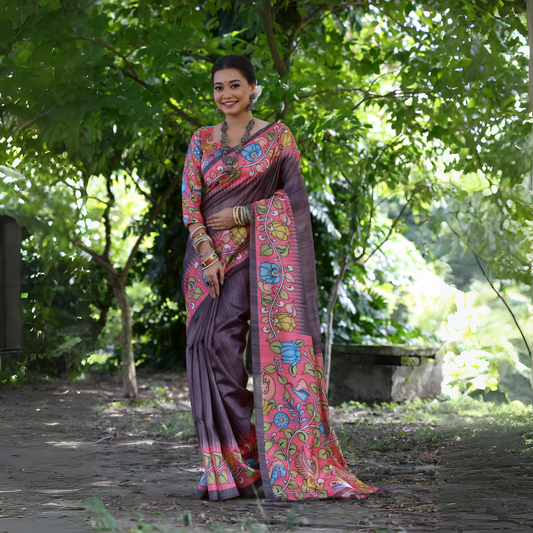 Woman wearing a mauve saree with vibrant Madhubani-inspired floral print border and pallu, paired with a matching blouse and traditional jewelry, standing gracefully on a garden pathway surrounded by lush green trees and foliage.