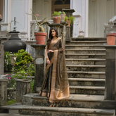 Woman wearing an olive green silk saree with a gold border and subtle woven motifs, accessorized with gold bangles, standing on the stone steps of a heritage mansion surrounded by potted plants and lush greenery