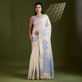 Woman wearing an ivory silk saree with blue floral motifs and a lavender blouse, accessorized with pearl jewelry, posing in a contemporary olive green studio with a geometric window shadow on the wall.
