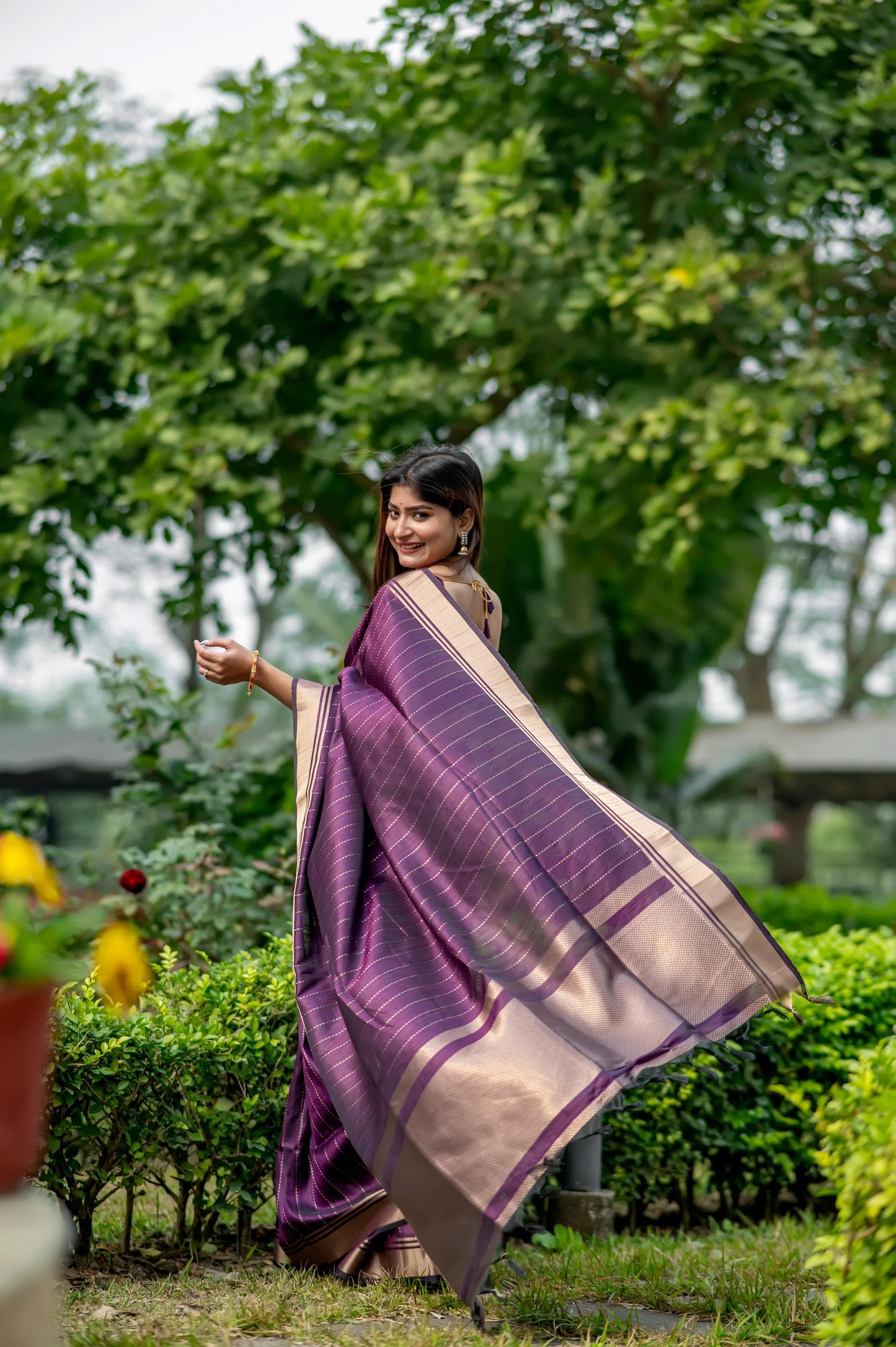 Woman in a deep shade purple saree with zari work posing gracefully in a garden with lush greenery and colorful flowers in the background