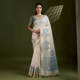 Woman wearing an ivory silk saree with grey fish and floral motifs, paired with a grey blouse and pearl jewelry, posing in a contemporary studio with olive green walls and a metal chair.