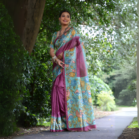 Woman wearing a mauve saree with vibrant Madhubani-inspired floral print border and pallu, paired with a matching blouse and traditional jewelry, standing gracefully on a garden pathway surrounded by lush green trees and foliage.