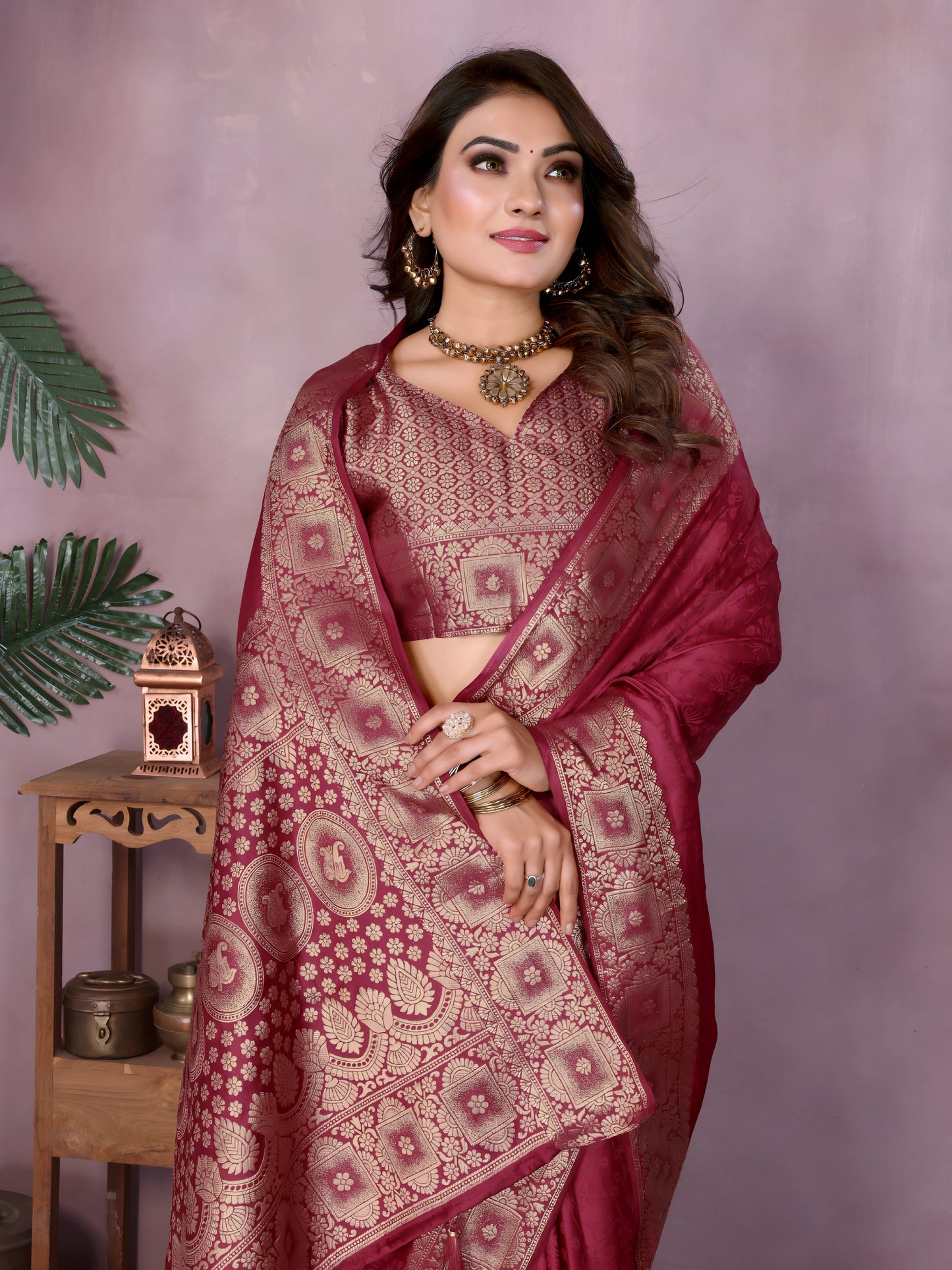 Woman wearing a mauve Banarasi silk saree adorned with an intricate golden zari border and detailed traditional motifs, paired with a matching blouse and gold jewelry. She stands gracefully in a modern studio featuring large rustic white clay pots, a wooden stand with brass decorative items, a bowl of colorful marigold flowers, and green foliage against a muted mauve backdrop.