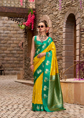 Woman in a yellow and green saree standing in front of a stone wall.