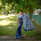 Woman wearing a blue handloom cotton saree with white tribal-inspired prints and a light blue blouse, accessorized with traditional jewelry, posing gracefully outdoors under lush green trees with sunlight filtering through the garden.