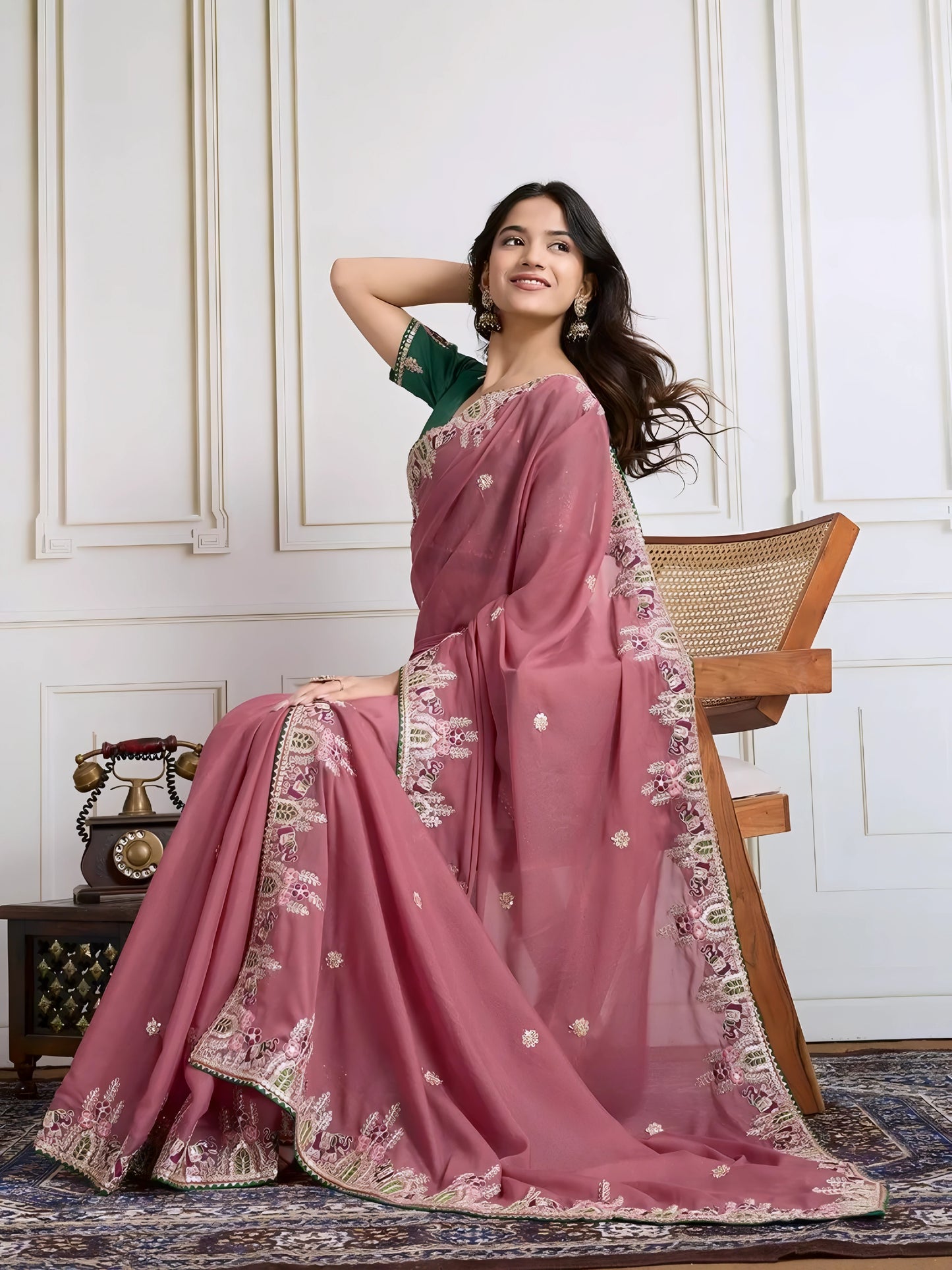 Woman wearing a dusty rose designer saree adorned with sequin detailing and an ornate embroidered floral border, paired with a contrasting green blouse embellished with small floral motifs. She stands elegantly in a classic white paneled room featuring a wooden armchair with a woven seat and a vintage rotary telephone on a carved wooden side table, all set atop an intricate patterned rug for a refined, traditional look.