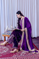 Woman in a purple traditional outfit sitting on a patterned rug with white curtains in the background.