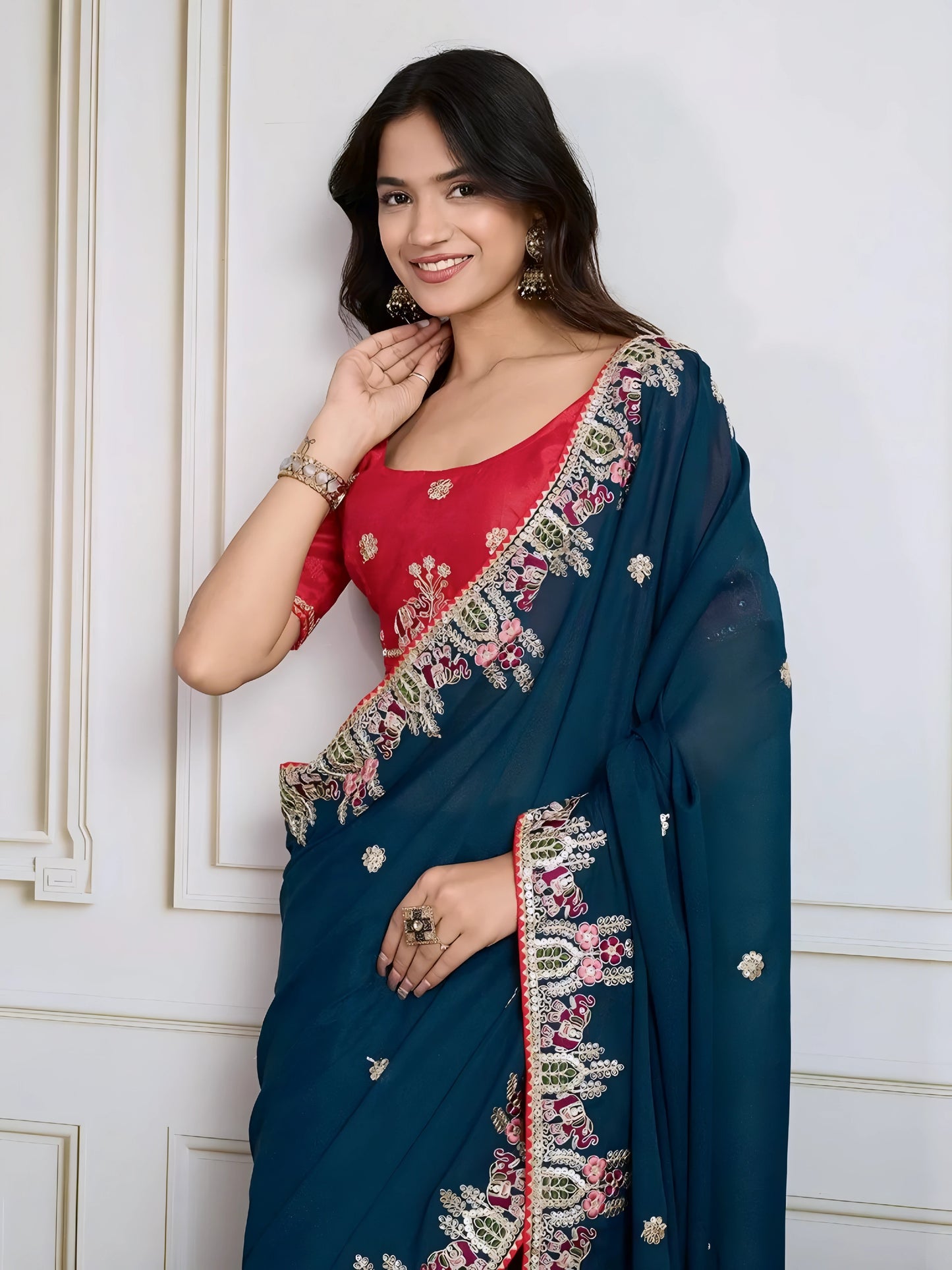 Woman wearing a teal blue designer saree embellished with beaded embroidery and ornate floral border, paired with a contrasting red blouse adorned with floral motifs. She poses stylishly in a classic white paneled room featuring a modern wooden armchair with a woven seat and a vintage rotary telephone on a carved side table, all set atop a patterned rug for an elegant, traditional look.