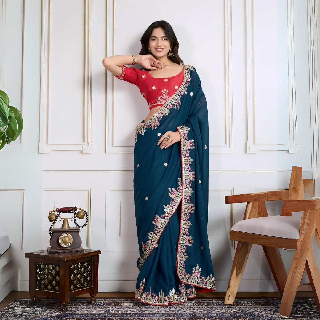 Woman wearing a teal blue designer saree embellished with beaded embroidery and ornate floral border, paired with a contrasting red blouse adorned with floral motifs. She poses stylishly in a classic white paneled room featuring a modern wooden armchair with a woven seat and a vintage rotary telephone on a carved side table, all set atop a patterned rug for an elegant, traditional look.