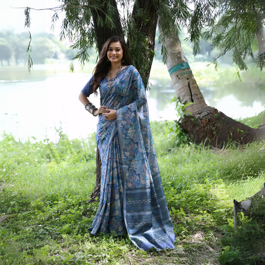 A woman models a Soft Tussar Silk Saree in various shades of blue, covered in an elaborate all-over artistic print. The pallu features dense blue and silver Zari weaving. She wears a blue blouse and a substantial set of oxidized silver jewelry, posing in a natural, outdoor setting.