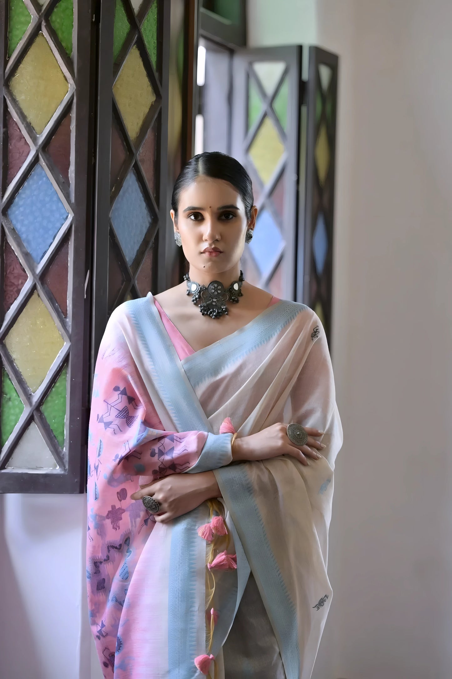 Woman wearing a white silk saree with pastel pink and blue village motifs and a tassel border, paired with a pink and blue blouse and statement necklace, standing indoors beside vintage stained glass windows