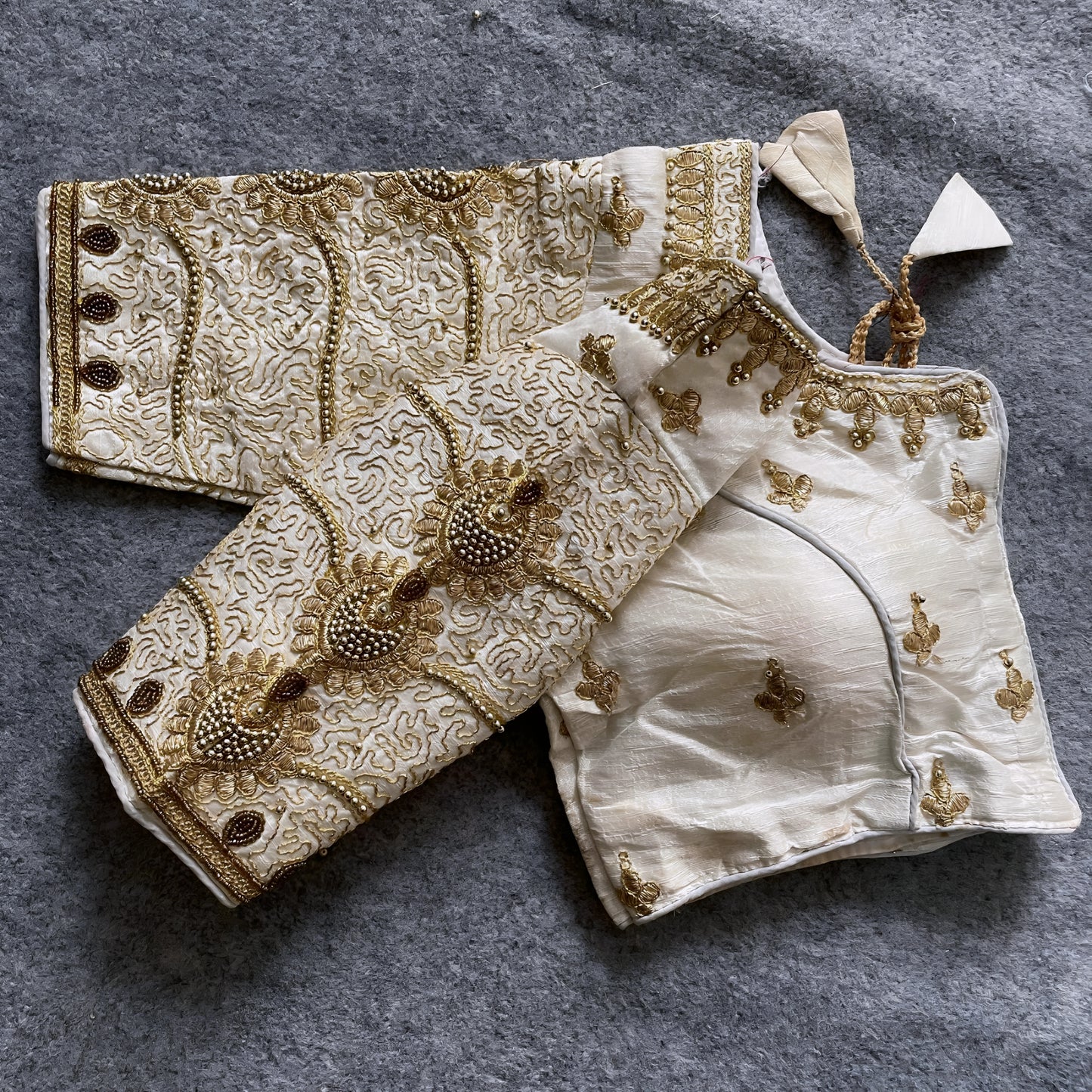 Traditional ivory silk blouse featuring intricate gold embroidery and beadwork with floral and paisley motifs on the sleeves and neckline, displayed flat on a textured surface with a bunch of artificial flowers and a woven mat in the background.