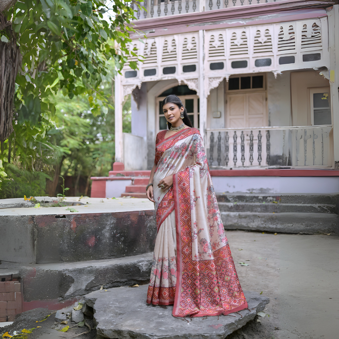 Woman wearing an off-white silk saree with red Patola-inspired geometric and floral borders, paired with a matching blouse and traditional jewelry, elegantly posing on stone steps in front of a vintage heritage house with ornate wooden railings and lush green trees in the background.