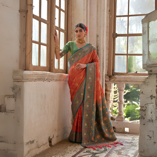 A woman models a vibrant Coral Orange Banarasi Silk Saree with all-over gold Zari weaving. The saree has a wide, ornate Rama Blue/Teal border featuring traditional Zari weaving. She wears a light green blouse and heavy gold Kundan jewelry, posing by a large antique window.