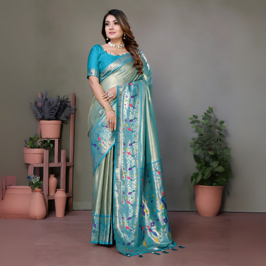 Woman wearing an elegant aqua blue silk saree featuring a pastel sheen, intricate floral and polka dot motifs in multicolor threads, and a matching blue blouse with silverwork on the sleeves. She is accessorized with a choker necklace, bangles, and rings, posing gracefully in a contemporary studio with minimalist decor, including potted green plants and coordinated terracotta vases on wooden stands against a muted backdrop.
