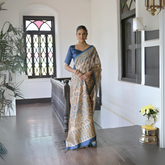Woman wearing a beige silk saree with intricate blue and gold geometric prints and a blue blouse, accessorized with a choker necklace and bangles, standing in a sunlit vintage hallway with wooden flooring, black railing, and stained glass windows.