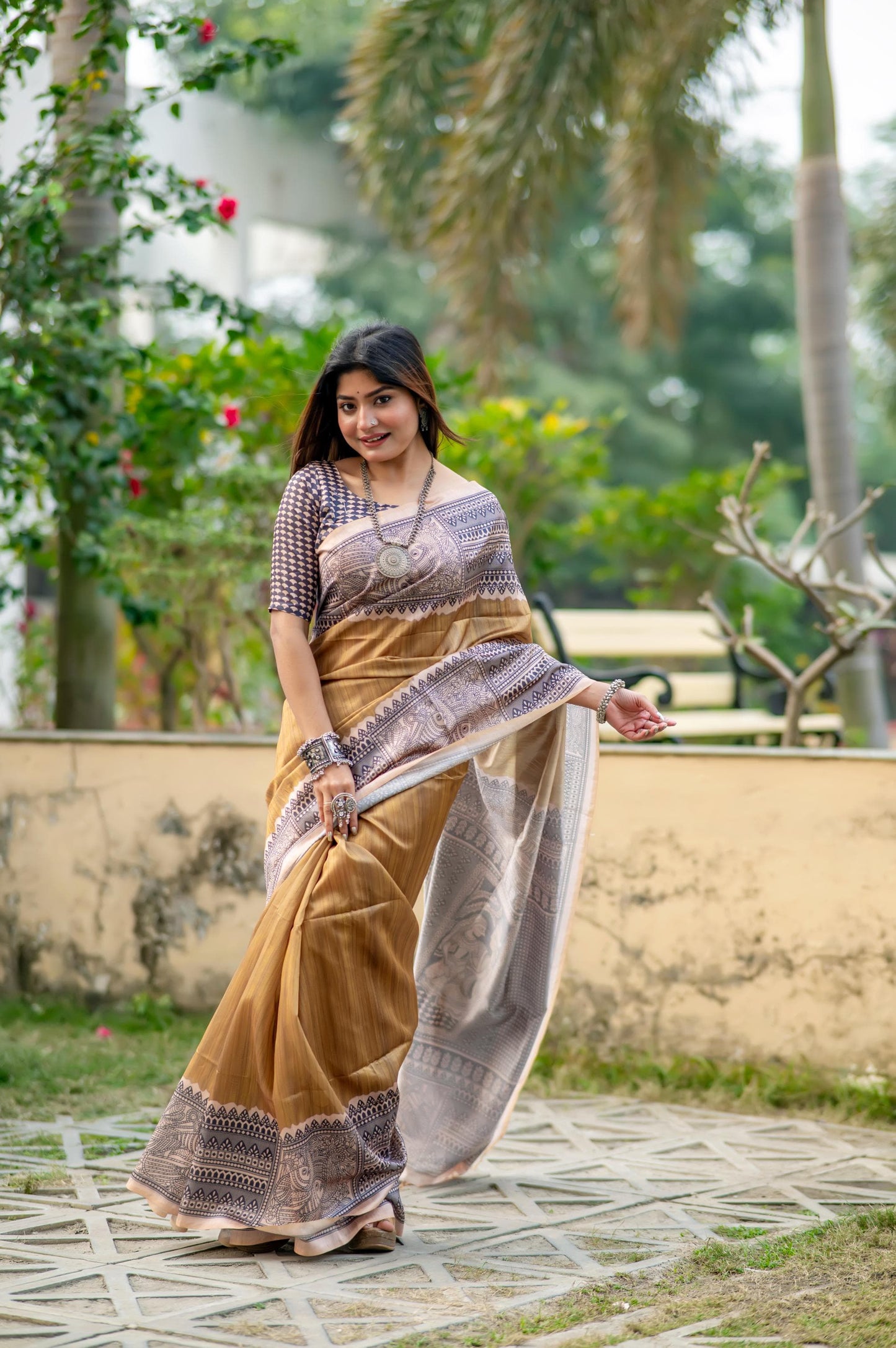 Woman in a beige saree with Madhubani print standing near a pond with lotus flowers in a lush garden setting