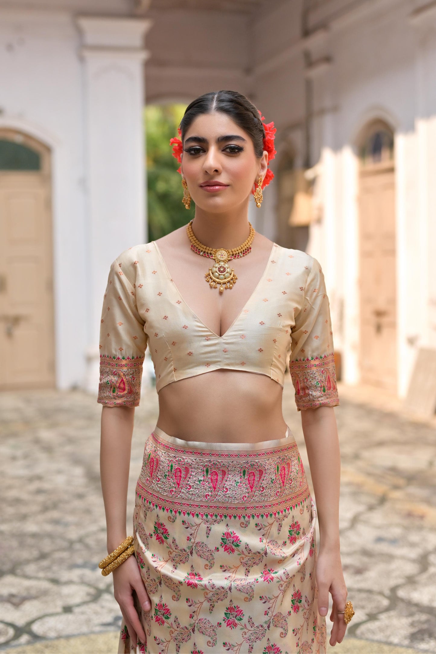 Woman in beige zari work saree with intricate patterns standing in a traditional courtyard
