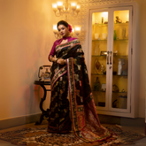 Woman wearing a black Banarasi silk saree with intricate multicolor floral zari work and a magenta blouse, accessorized with gold jewelry, standing in a warmly lit vintage interior featuring a chandelier, antique telephone, and a glass display cabinet.