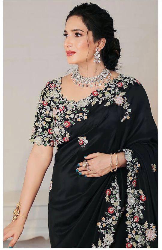 Woman wearing a black saree with intricate embroidery work, accessorized with a necklace and earrings, standing in a room with wooden flooring