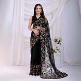 Woman wearing a black net saree with vibrant multicolor floral embroidery and a matching sleeveless blouse, posing in a modern white studio with draped curtains and white floral arrangements in the background.