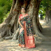 Woman wearing a black silk saree with vibrant multicolor floral motifs and a contrasting coral pink border, paired with a matching blouse and traditional jewelry, elegantly standing in front of a large, ancient tree with exposed roots in a serene outdoor setting.