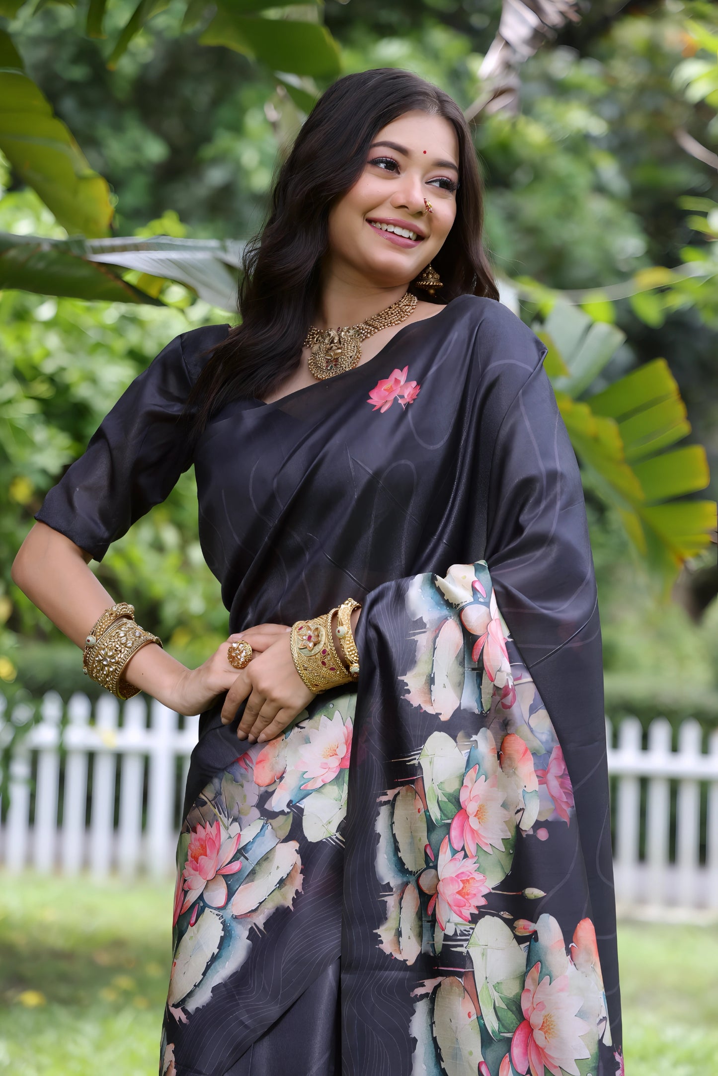 Woman wearing a black saree with large handpainted lotus and leaf floral motifs, accessorized with traditional jewelry, posing gracefully outdoors in a lush green garden with tropical plants and a white picket fence in the background.