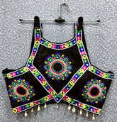 Black blouse featuring vibrant circular and oval mirror work with traditional Kutch multicolor embroidery, adorned with bold geometric borders and shell tassel drops along the hem. Displayed on a hanger against a textured silver background, highlighting its festive and culturally inspired ethnic design.