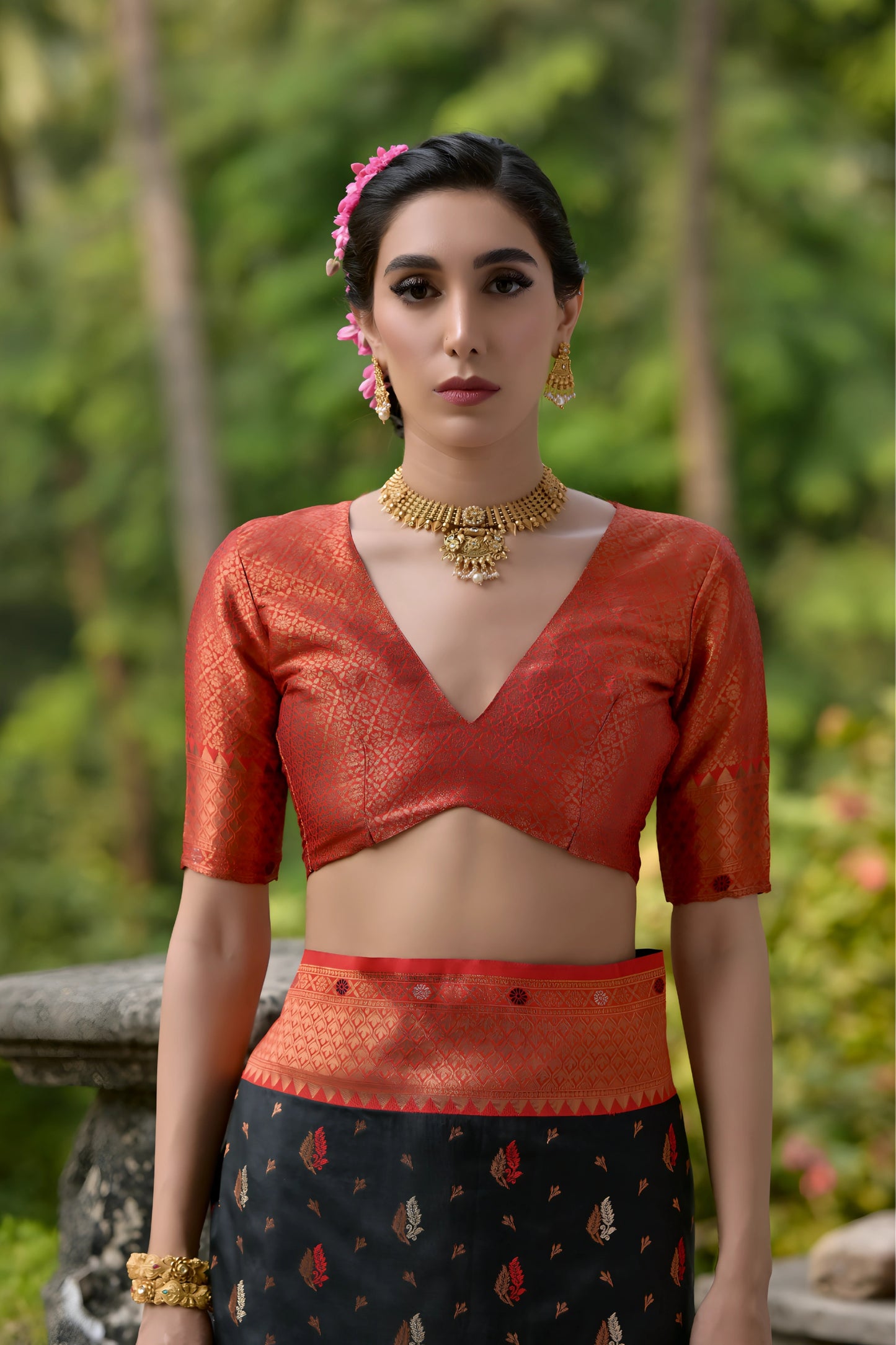Woman wearing a black and orange silk saree with gold motifs and a contrasting orange blouse, posing outdoors on a veranda with lush green trees in the background.