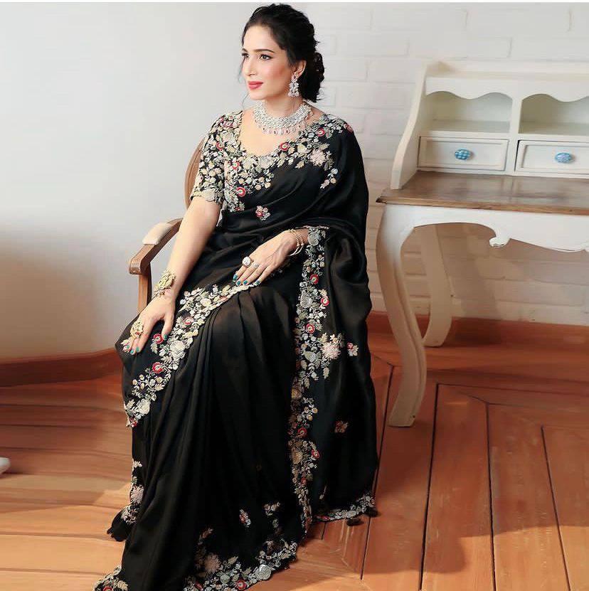 A person wearing a black saree adorned with intricate embroidery work, standing in a room with wooden flooring and light-colored curtains.