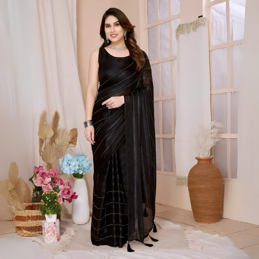 Woman wearing a black saree with subtle sequin stripes and a sleeveless blouse, accessorized with silver jewelry, standing in a modern interior with large windows, vases of pink and blue flowers, and neutral decor elements.