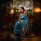 Woman wearing a blue Banarasi silk saree with intricate gold zari motifs and border, paired with a purple blouse and traditional jewelry, seated in a vintage-inspired interior featuring a gramophone, checkered flooring, and warm ambient lighting.