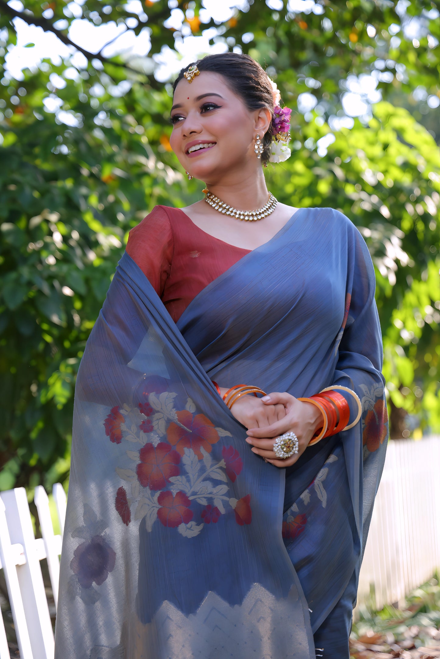 Woman wearing a blue silk saree with red floral prints and a contrasting red blouse, accessorized with pearl jewelry and orange bangles, standing beside a white picket fence in a sunlit garden with lush green trees in the background.