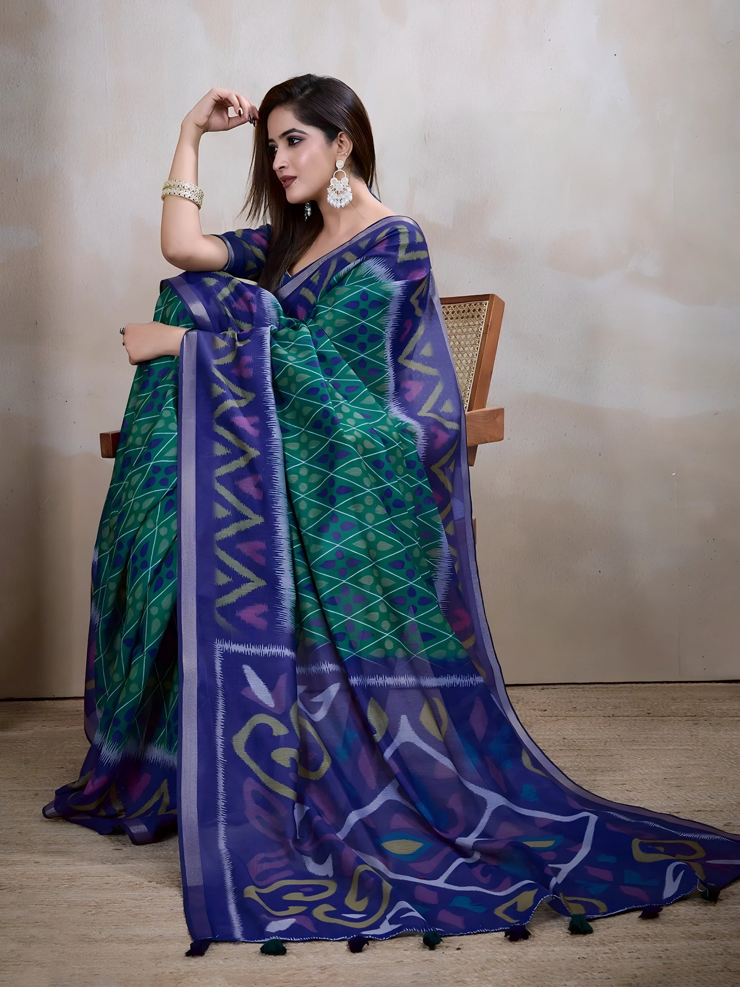 Woman wearing a blue and green handloom silk saree featuring bold geometric patterns, zigzag lines, and diamond motifs, paired with a matching blue blouse. She is styled with silver jewelry and bangles, posing gracefully in a contemporary studio setting with a neutral wall, a cane partition, a decorative lamp, and minimal furnishings for an elegant traditional look.