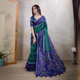 Woman wearing a blue and green handloom silk saree featuring bold geometric patterns, zigzag lines, and diamond motifs, paired with a matching blue blouse. She is styled with silver jewelry and bangles, posing gracefully in a contemporary studio setting with a neutral wall, a cane partition, a decorative lamp, and minimal furnishings for an elegant traditional look.