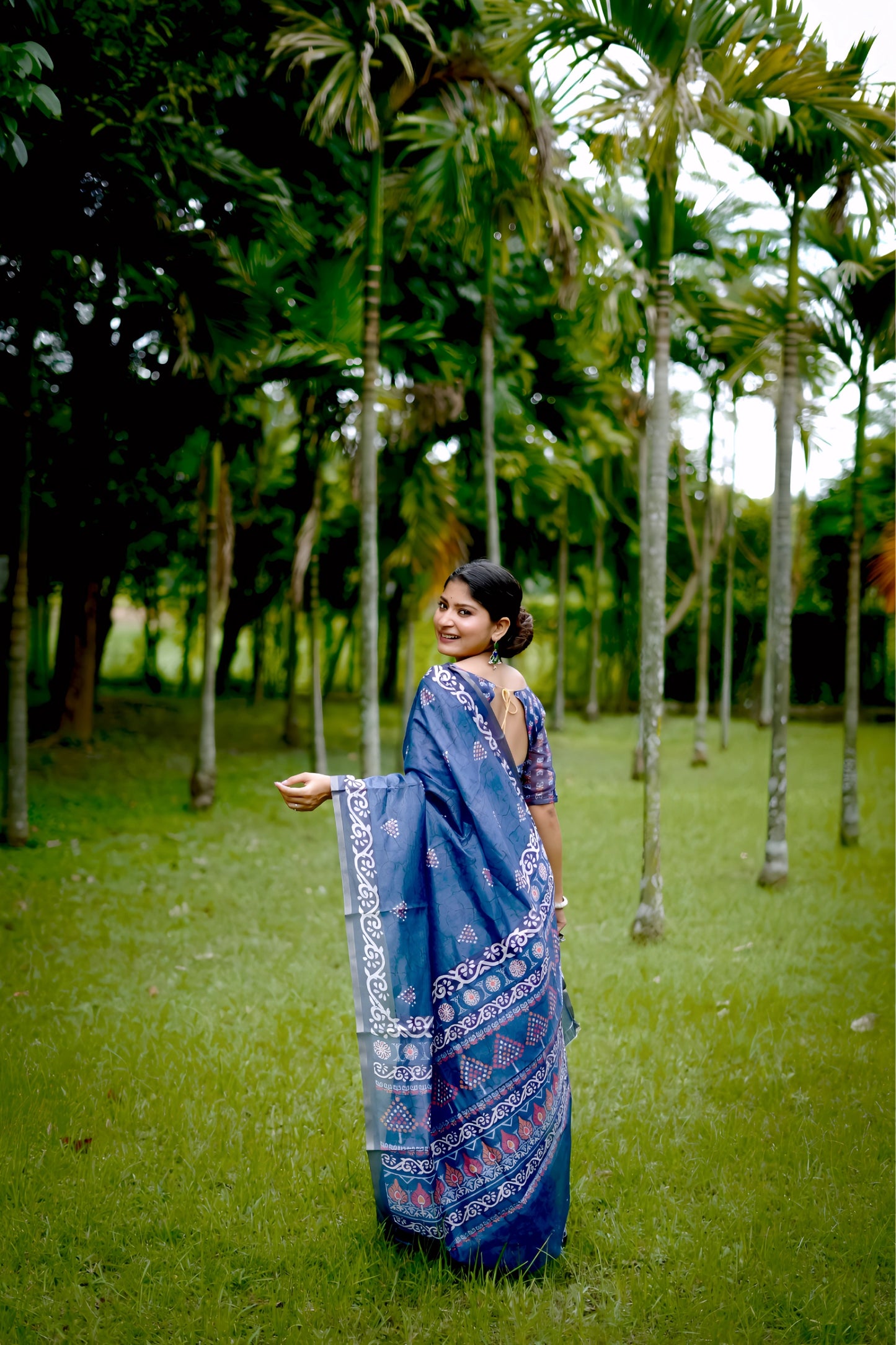 Woman wearing a blue handloom cotton saree with white tribal-inspired border and geometric patterns, paired with a matching printed blouse and silver jewelry, standing gracefully in a lush green garden with tall trees in the background.