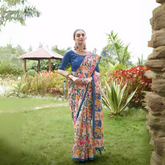 Woman wearing a blue blouse and a vibrant Madhubani-printed saree with multicolor floral and animal motifs, accessorized with traditional jewelry, standing gracefully on a lush green lawn surrounded by tropical plants and red foliage, with a garden gazebo in the background.