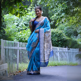Woman wearing a blue Madhubani-printed saree with an intricate white border and traditional motifs, paired with a matching blue blouse and gold jewelry, standing gracefully on a garden path lined with a white picket fence and surrounded by lush green foliage.