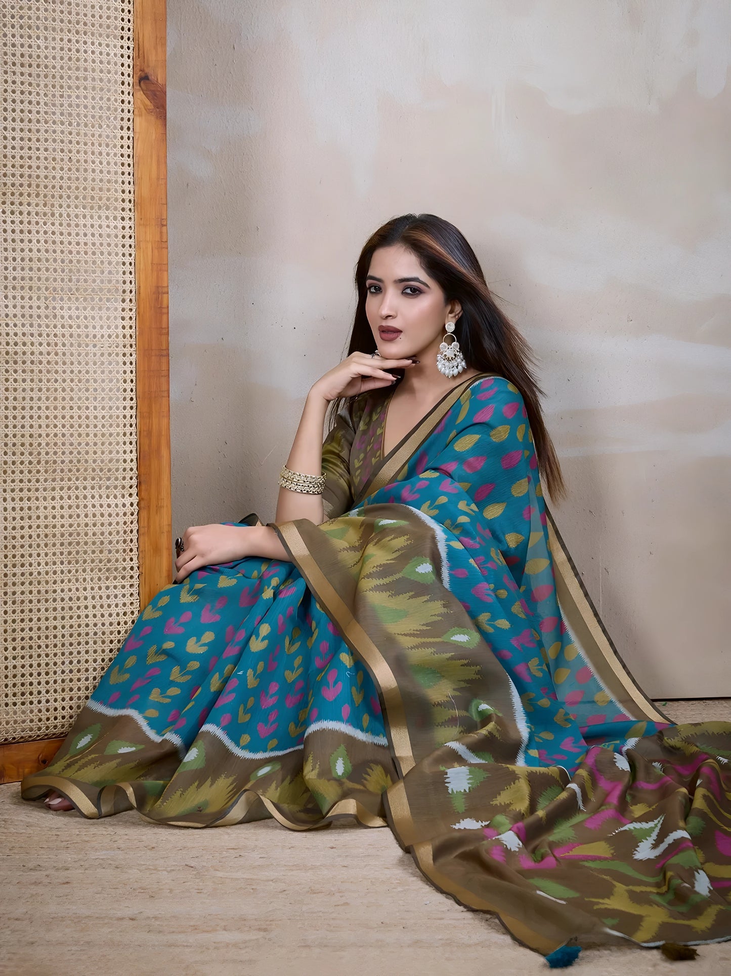 Woman wearing a blue silk saree featuring vibrant pink and olive green ikat-inspired abstract patterns, paired with a matching blouse and elegant gold bangles. She poses gracefully in a contemporary studio setting with a neutral textured wall, a tall woven cane screen, and rustic wooden flooring, highlighting the saree’s flowing pallu and artistic design.