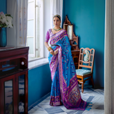 Woman wearing a blue Patola silk saree with purple and gold borders, paired with a purple blouse and traditional jewelry, standing gracefully by a window in a vintage-inspired room with teal blue walls, wooden furniture, and floral decor.