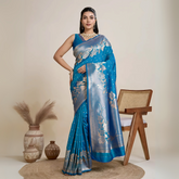 Woman wearing a vibrant blue silk saree with silver floral zari border and pallu, paired with a matching blouse and traditional jewelry, elegantly posing beside a wooden chair in a minimalist studio with neutral decor and ceramic vases.