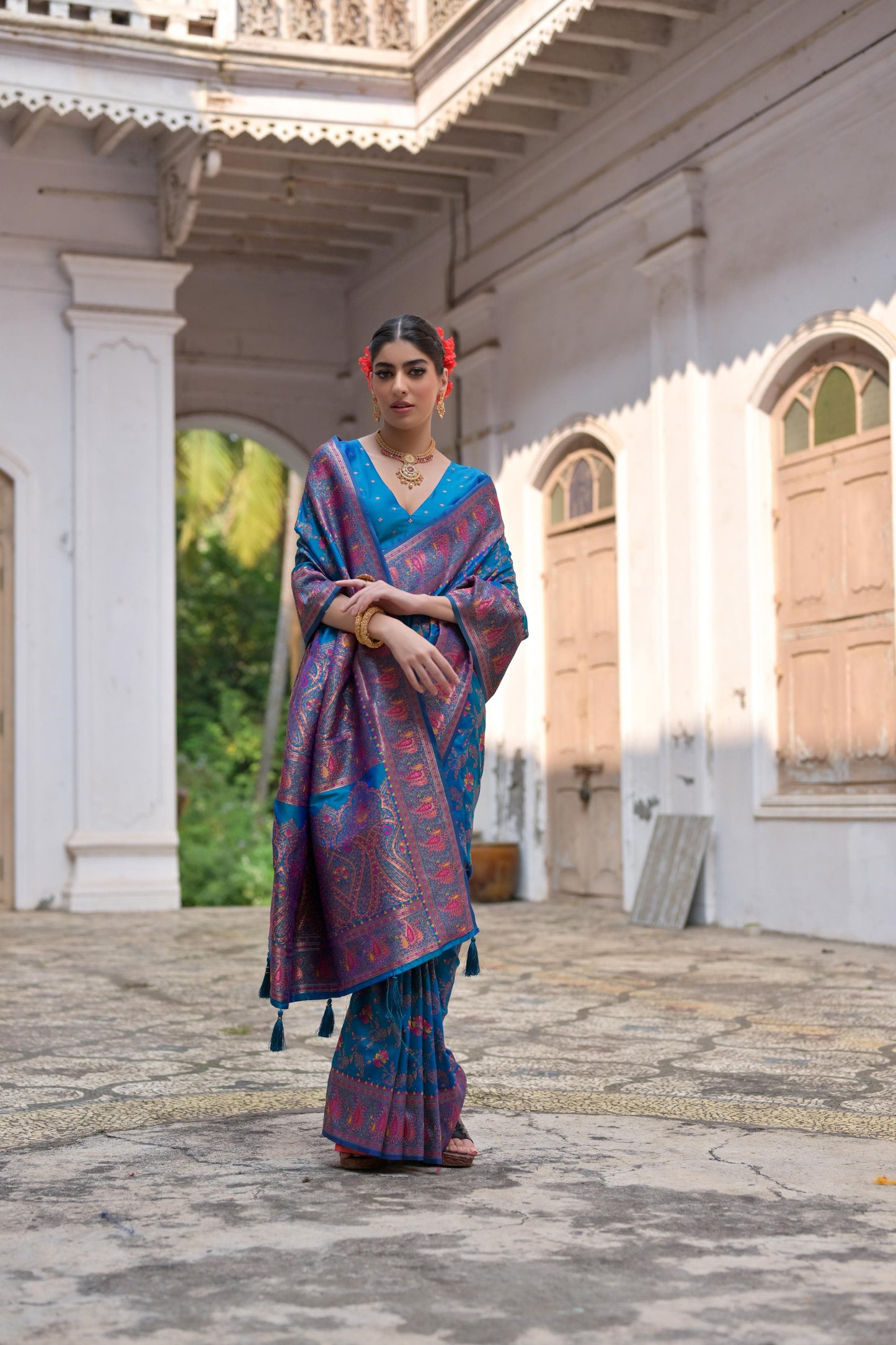 Woman in blue zari work saree with intricate patterns standing in a traditional courtyard