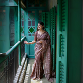 Woman wearing a brown floral printed saree with a matching blouse and statement earrings, standing on a vintage balcony with teal green shutters and doors, bathed in natural light.