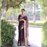 Woman wearing a brown saree with gold border and floral embroidery, posing outdoors under a leafy tree with lush greenery and palm trees in the background.
