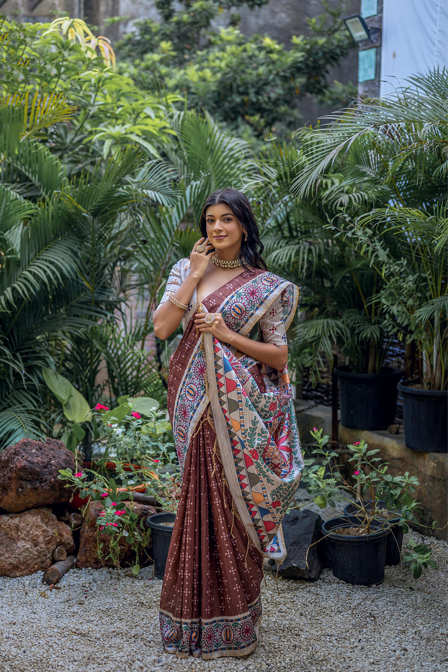 Woman wearing a brown saree with intricate white tribal-inspired prints and a multicolor pallu featuring circular and geometric patterns, accessorized with traditional jewelry, standing gracefully in a lush outdoor garden surrounded by tropical plants.