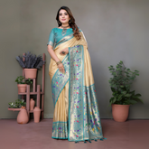 Woman wearing a cream silk saree with a sky blue border and multicolor floral motifs, paired with a matching blue blouse and traditional jewelry. She poses gracefully in a modern studio with minimalist decor, featuring potted plants and terracotta vases on wooden stands against a muted backdrop.