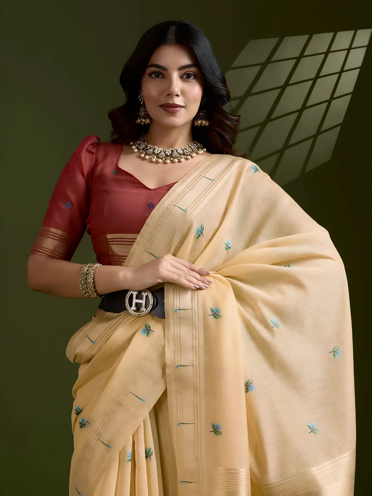 Woman wearing a cream silk saree adorned with delicate floral embroidery and tassel detailing on the pallu, paired with a red blouse and traditional jewelry, elegantly posing in a contemporary studio with olive green walls and decorative window light patterns.