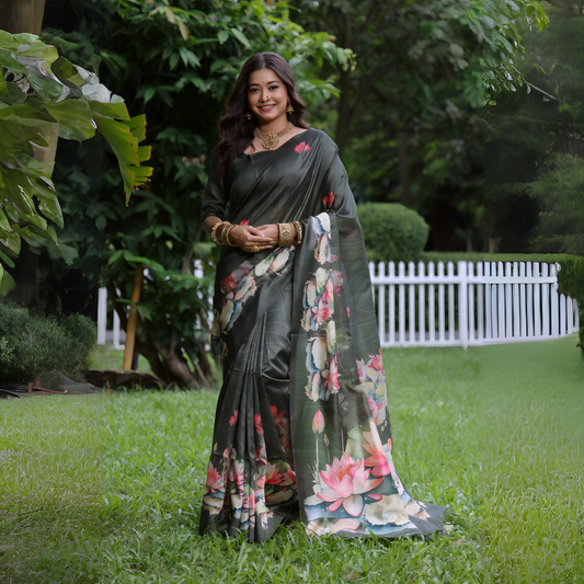 Woman wearing a dark green saree with large handpainted lotus and leaf floral motifs, accessorized with traditional gold jewelry and bangles, standing on a lush green lawn with tropical plants and a white picket fence in the background