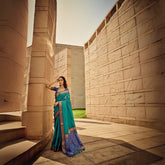 Graceful woman showcasing deep green saree with intricate zari work and detailed pallu, standing by modern sandstone structures under sunny sky